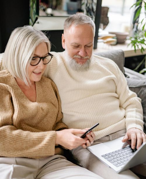 Senior couple using smartphone and laptop together on a cozy sofa indoors, depicting modern technology lifestyle.