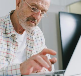 Elderly man in plaid shirt using a laptop indoors.